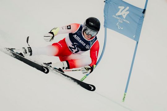 Ein Skifahrer in rot-weißer Kleidung rast um eine blaue Paralympics-Flagge von Milano Cortina 2026 herum bergab, wobei er sich stark neigt und Schnee aufwirbelt, während er einen Helm und eine Brille trägt.