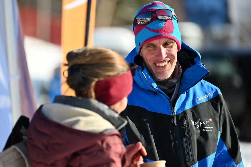 Ein Mann in blauer Winterjacke und bunter Mütze lächelt, während er an einem sonnigen Wintertag im Freien mit einer Frau mit kastanienbraunem Stirnband und Jacke spricht. Der Mann hält eine Tasse in der Hand und trägt eine Sonnenbrille auf seiner Mütze.