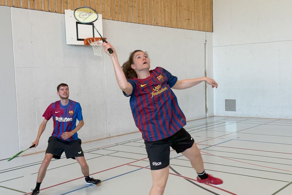 Zwei Personen in passenden Trikots des FC Barcelona spielen in einer Halle Badminton. Die Frau im Vordergrund springt, um mit ihrem Schläger den Federball zu treffen, während der Mann hinter ihr zusieht und bereit ist zu spielen.