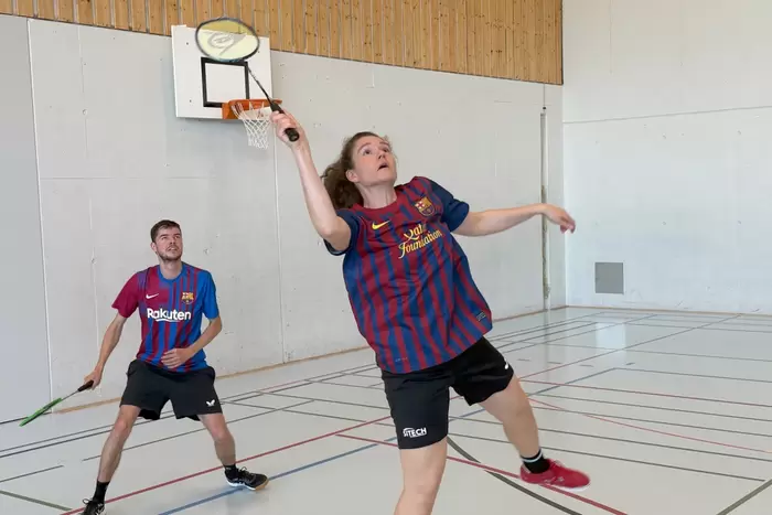 Zwei Personen in passenden Trikots des FC Barcelona spielen in einer Halle Badminton. Die Frau im Vordergrund springt, um mit ihrem Schläger den Federball zu treffen, während der Mann hinter ihr zusieht und bereit ist zu spielen.