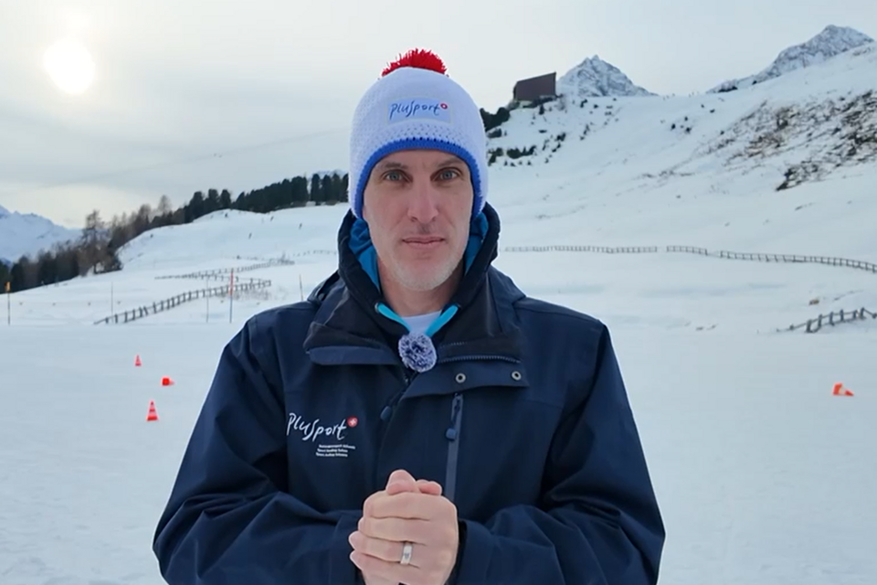 Ein Mann in dunkelblauer Jacke und weißer Wintermütze mit rotem Bommel steht in einer verschneiten Landschaft mit Bergen im Hintergrund, die Hände vor sich verschränkt, und schaut in die Kamera.