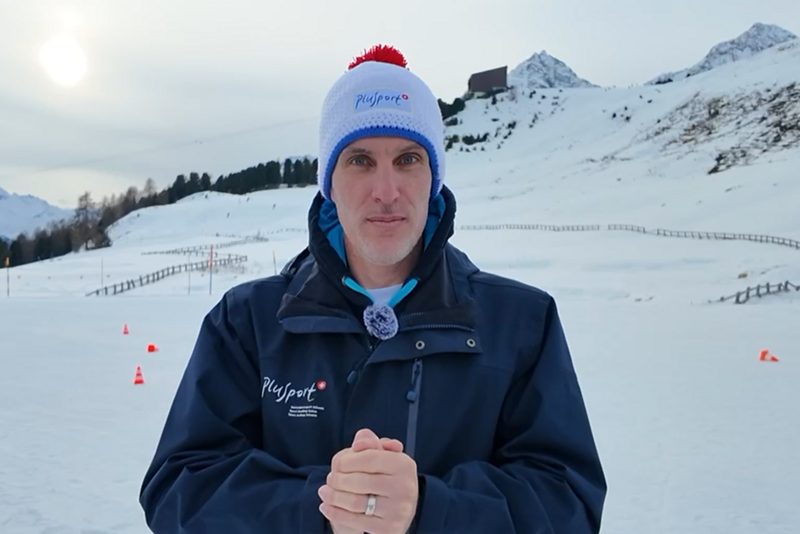 Ein Mann in dunkelblauer Jacke und weißer Wintermütze mit rotem Bommel steht in einer verschneiten Landschaft mit Bergen im Hintergrund, die Hände vor sich verschränkt, und schaut in die Kamera.
