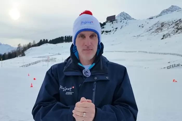 Ein Mann in dunkelblauer Jacke und weißer Wintermütze mit rotem Bommel steht in einer verschneiten Landschaft mit Bergen im Hintergrund, die Hände vor sich verschränkt, und schaut in die Kamera.