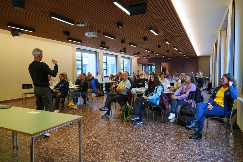 Ein Mann steht und spricht zu einem sitzenden Publikum in einem modernen, gut beleuchteten Raum mit großen Fenstern und einer holzgetäfelten Decke. Die Zuhörer hören aufmerksam zu, einige machen sich Notizen, während Tische und Taschen zu sehen sind.