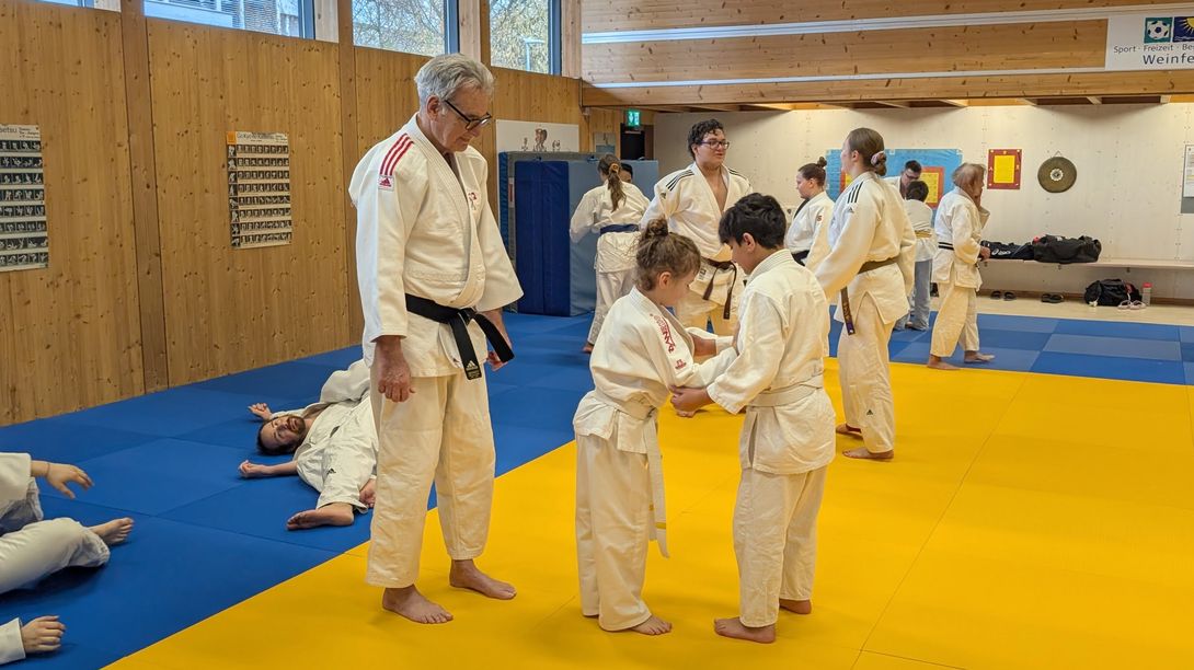 Eine Gruppe von Personen in weißen Judoanzügen trainiert in einem Dojo mit gelben und blauen Matten. Ein Ausbilder beobachtet zwei Kinder, die sich gegenüberstehen, während andere im Hintergrund trainieren oder sich ausruhen.
