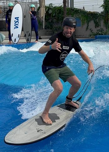 Ein Mann in einem schwarzen T-Shirt und grünen Shorts surft drinnen auf einer künstlichen Welle, lächelt und zeigt den Daumen nach oben. Im Hintergrund stehen zwei Personen mit Helmen und Surfbrettern.