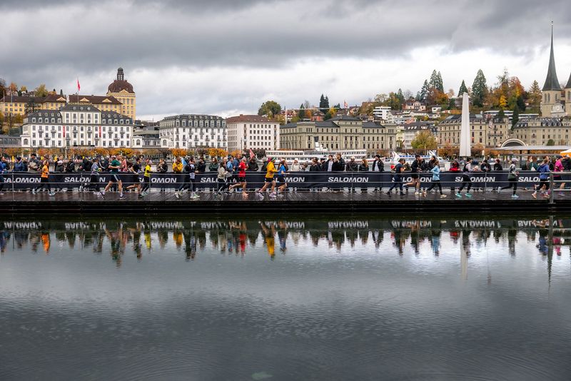 Läufer überqueren eine Brücke während eines Rennens in einer europäischen Stadt mit historischen Gebäuden im Hintergrund, bewölktem Himmel darüber und der Stadtlandschaft, die sich im ruhigen Wasser darunter spiegelt.
