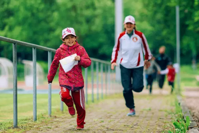 Ein junges Mädchen in roter Jacke und Mütze läuft lächelnd und mit einem Blatt Papier in der Hand über einen Weg im Freien, während ein Erwachsener und andere Personen hinter ihr herlaufen. Die Szene ist lebhaft und von Grün umgeben.