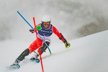 Ein Skifahrer in roter Rennkleidung und mit weißem Helm fährt einen Slalomkurs auf einer verschneiten Piste, wobei er sich stark um eine Stange lehnt, während im Hintergrund Schnee fällt.