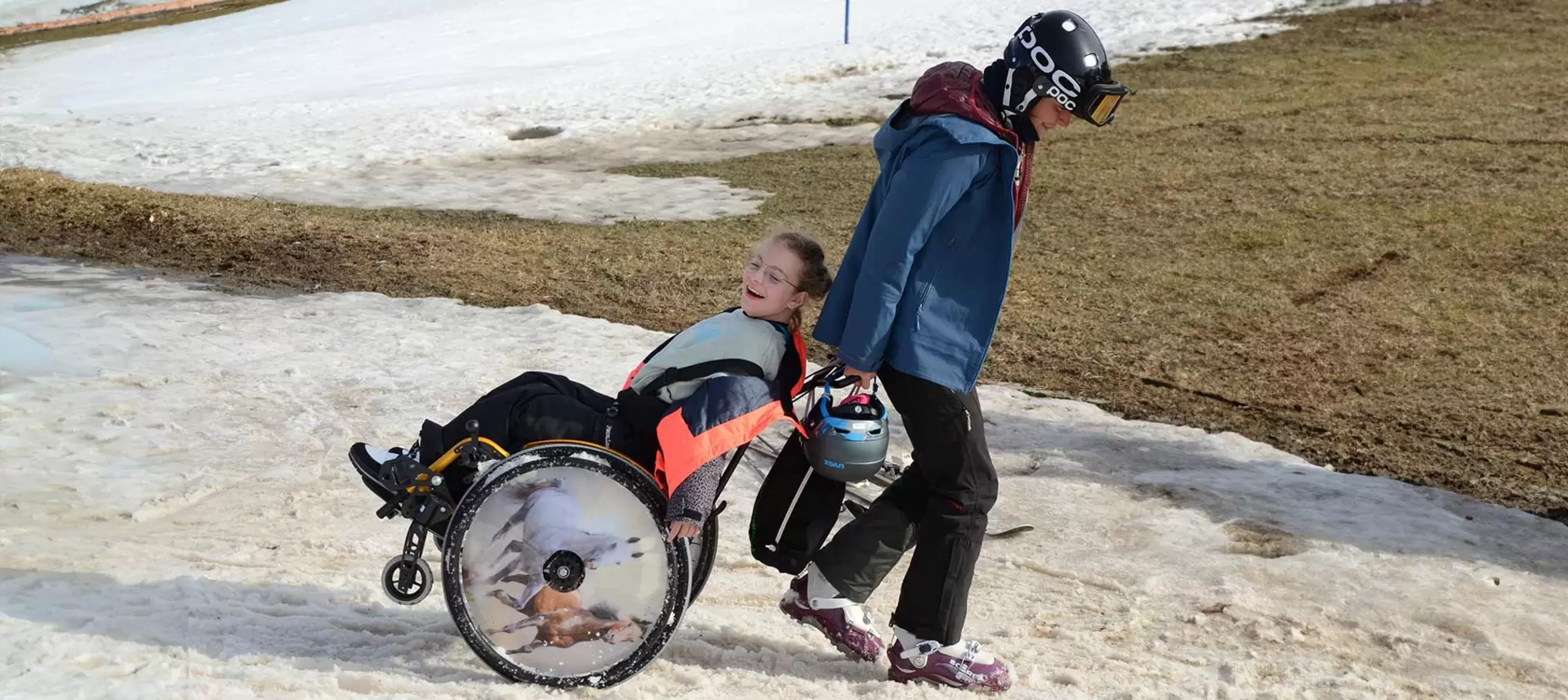 Eine Teilnehmerin im Rollstuhl wird von der Camps-Leiterin über ein Schneefeld gezogen. Beide lachen.