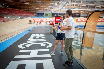 Zwei Personen stehen und unterhalten sich neben einer Indoor-Radrennbahn, während im Hintergrund Radfahrer fahren. Im Vordergrund sind ein großer GRENCHEN-Schriftzug auf dem Boden und ein geschwungenes orangefarbenes Banner zu sehen.