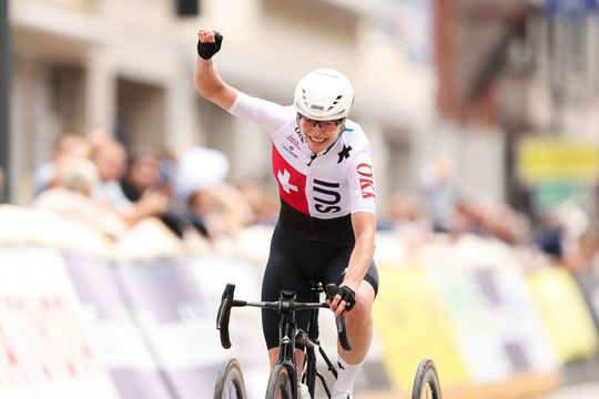 Ein Radfahrer, der einen weißen Helm und ein Trikot mit der SUI und einer Schweizer Flagge trägt, hebt beim Überqueren der Ziellinie während eines Rennens jubelnd die Faust. Der Hintergrund ist unscharf mit Zuschauern und Bannern.