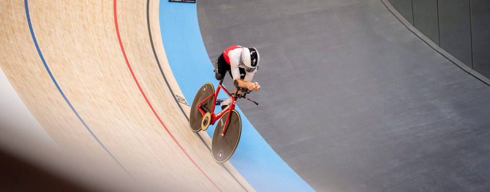 Ein Radfahrer, der einen Helm und eine Rennausrüstung trägt, fährt mit einem roten Fahrrad auf einer Indoor-Velodrombahn, wobei er sich in einen gebogenen Abschnitt mit blauen und hölzernen Fahrspuren hineinlehnt.