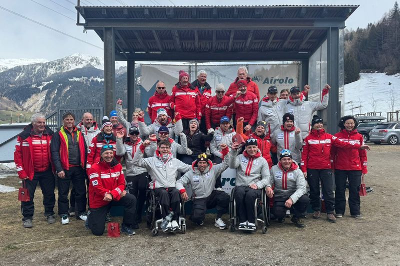Eine große Gruppe von Menschen, einige in Rollstühlen, posieren und jubeln gemeinsam im Freien in Winterkleidung. Im Hintergrund sind verschneite Berge und ein Gebäude zu sehen. Die meisten tragen passende rote oder graue Jacken.