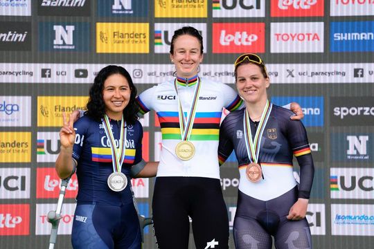 Drei Radfahrerinnen mit Medaillen stehen lächelnd auf einem Podium. Die mittlere Radfahrerin trägt ein Regenbogentrikot und eine Goldmedaille, flankiert von Silber- und Bronzemedaillengewinnerinnen. Im Hintergrund sind verschiedene Sponsorenlogos zu sehen.