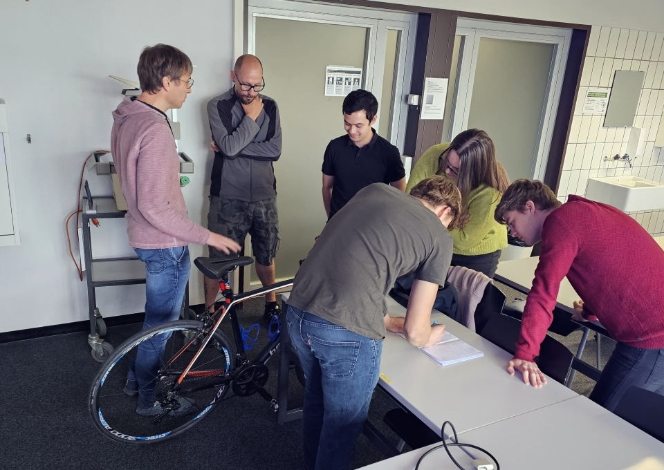 Sechs Personen sitzen in einem Raum um einen Tisch und unterhalten sich, während eine Person in ein Notizbuch schreibt. Neben ihnen steht ein Fahrrad. Einige Personen stehen, während andere sich über den Tisch beugen oder am Tisch sitzen.