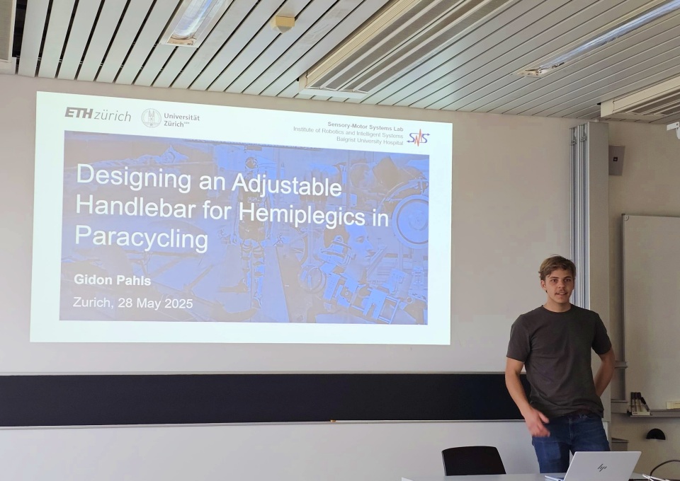 Ein junger Mann steht neben einer projizierten Präsentationsfolie mit dem Titel „Designing an Adjustable Handlebar for Hemiplegics in Paracycling“ von Gidon Pahls vom 28. Mai 2025 an der ETH Zürich.