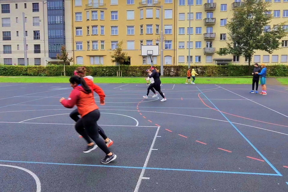 Menschen laufen auf einem Basketballplatz im Freien, im Hintergrund stehen oder gehen einige andere. Der Platz befindet sich an einem bewölkten Tag vor einem mehrstöckigen Wohnhaus.