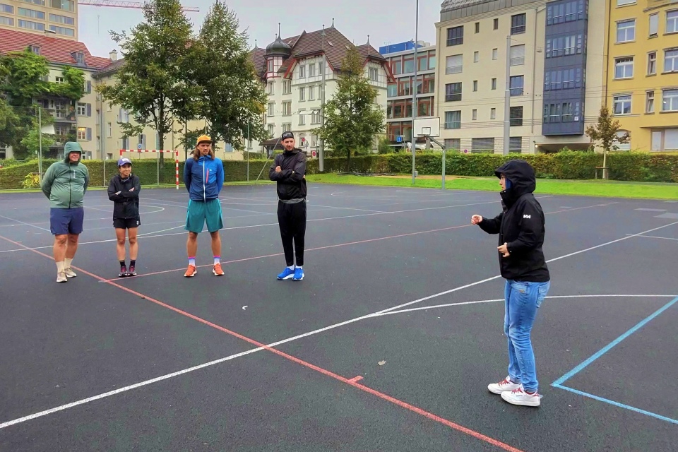 Fünf Personen stehen in einem Halbkreis auf einem Basketballplatz im Freien, bekleidet mit Jacken und Kapuzenpullis; eine andere Person steht ihnen gegenüber und spricht oder gibt Anweisungen. Im Hintergrund sind Gebäude und Bäume unter einem bedeckten Himmel zu sehen.