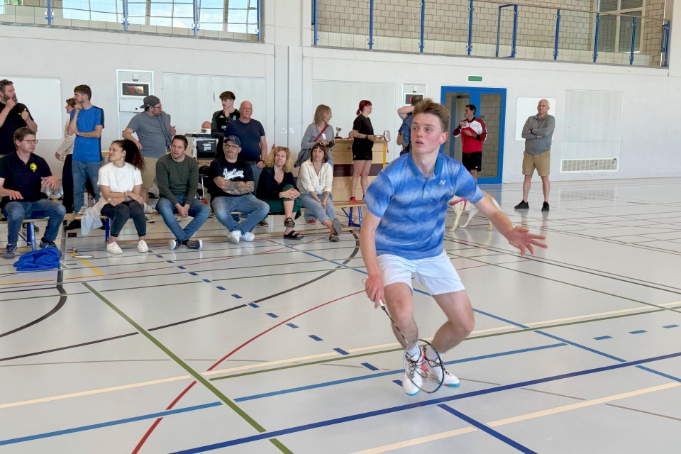 Ein junger Mann in einem blauen Hemd und weißen Shorts spielt Badminton in einer Turnhalle. Er konzentriert sich auf das Spiel und hält einen Schläger in der Hand, während im Hintergrund Zuschauer sitzen und zusehen.