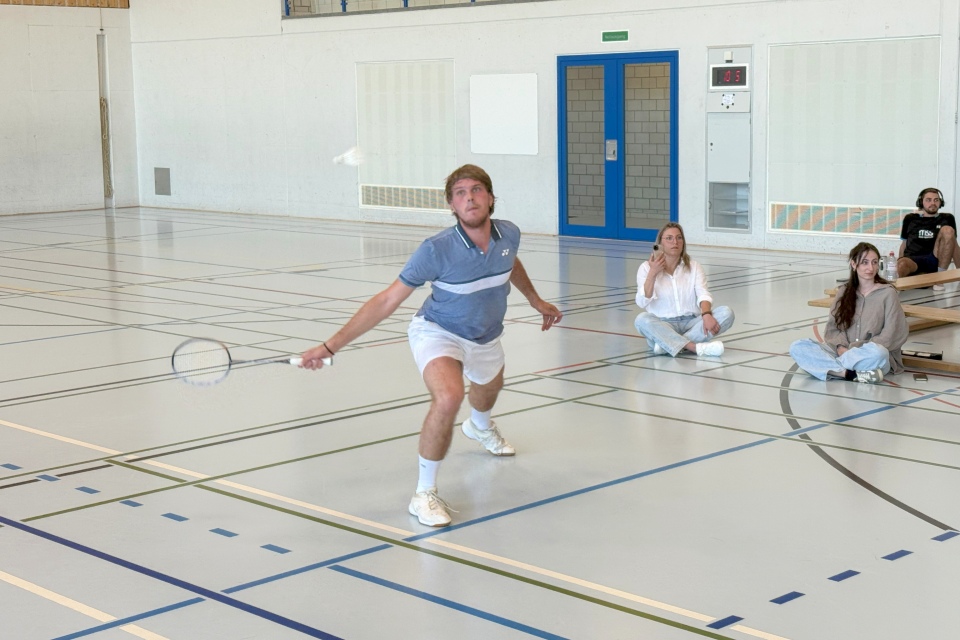 Ein Mann in Sportkleidung macht einen Ausfallschritt, um einen Federball mit einem Badmintonschläger in einer Turnhalle zu schlagen, während drei Personen auf Bänken im Hintergrund sitzen und das Spiel beobachten.