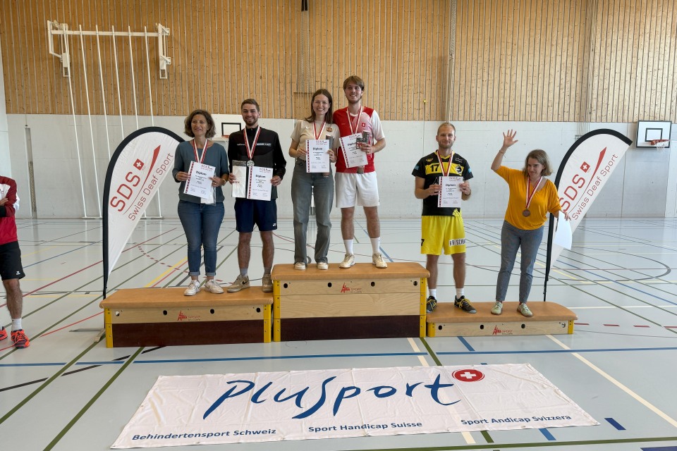 Sechs Personen stehen auf einem Podium in einer Turnhalle, halten Urkunden und tragen Medaillen. Zwei Transparente und ein Plusport-Banner sind zu sehen. Die Gruppe lächelt und freut sich über eine sportliche Leistung.