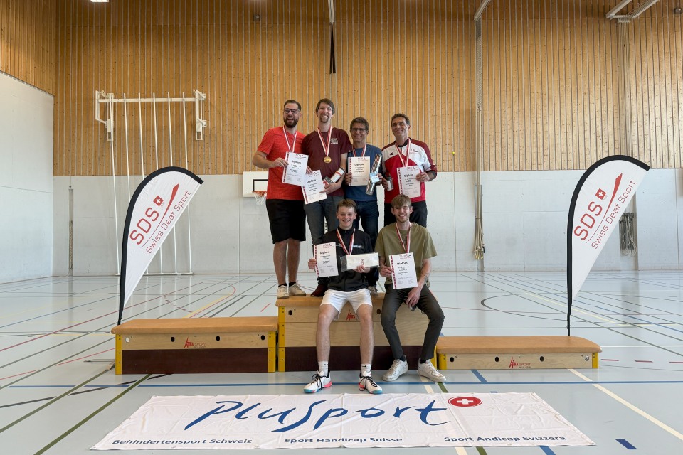 Sechs Männer posieren auf einem Podium in einer Turnhalle, halten Urkunden und tragen Medaillen. Zwei Banner und ein Plusport-Schild sind zu sehen, die auf eine Sportveranstaltung hinweisen. Im Hintergrund sind eine Holzwand und ein Basketballkorb zu sehen.