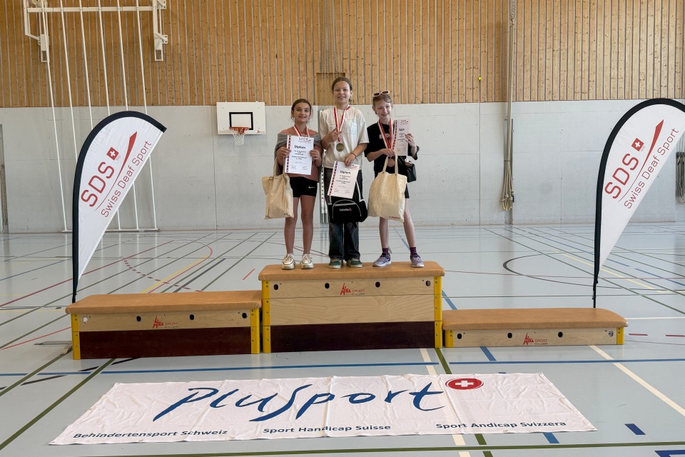 Drei junge Mädchen stehen auf einem Podium in einer Turnhalle und halten Urkunden und Medaillen in der Hand. Zwei SDS-Fahnen und ein Plusport-Banner weisen auf eine Sportveranstaltung oder einen Wettbewerb für Jugendliche hin.