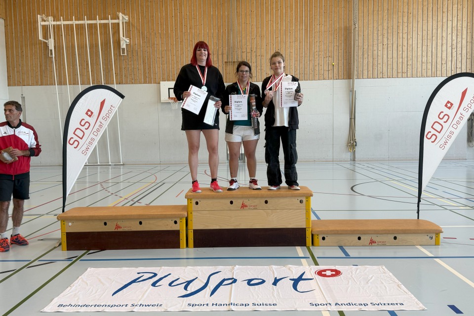 Drei Frauen stehen auf einem Podium mit Urkunden und Medaillen in einer Sporthalle. Zwei Banner mit dem SDS- und dem PluSport-Logo flankieren das Podium, davor hängt ein großes PluSport-Banner auf dem Boden.
