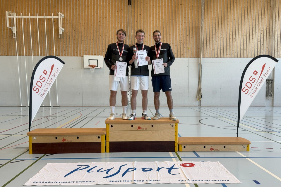 Drei männliche Athleten stehen auf einem Podium mit Urkunden und Medaillen in einer Sporthalle. Zwei Fahnen mit SDS flankieren das Podium, und auf dem Boden davor ist ein Plusport-Banner angebracht.