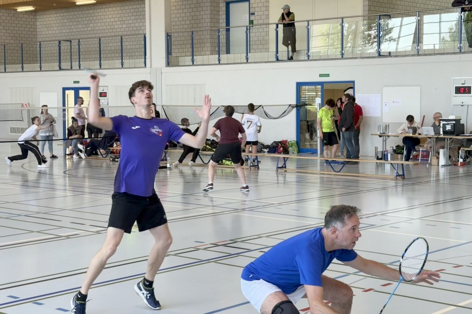 Zwei Männer spielen Badminton in einer Sporthalle; der eine im lila Hemd bereitet sich darauf vor, den Federball zu schlagen, während sein Mannschaftskamerad in Blau hockt. Im Hintergrund der hell erleuchteten Sporthalle laufen Menschen herum und schauen zu.