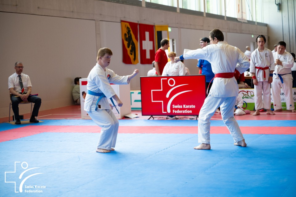 Zwei Karatesportler stehen sich während eines Wettkampfs auf einer blauen und roten Matte gegenüber. Der eine trägt einen blauen, der andere einen roten Gürtel. Kampfrichter und Zuschauer beobachten, im Hintergrund ist ein Schild des Schweizerischen Karateverbandes zu sehen.