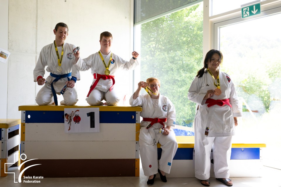 Vier Karate-Athleten posieren mit Medaillen auf einem Podest in der Halle und feiern ihre Erfolge. Drei tragen weiße Uniformen mit blauen oder roten Gürteln, einer trägt schwarze Sandalen. In der Ecke ist das Logo des Schweizerischen Karateverbandes zu sehen.