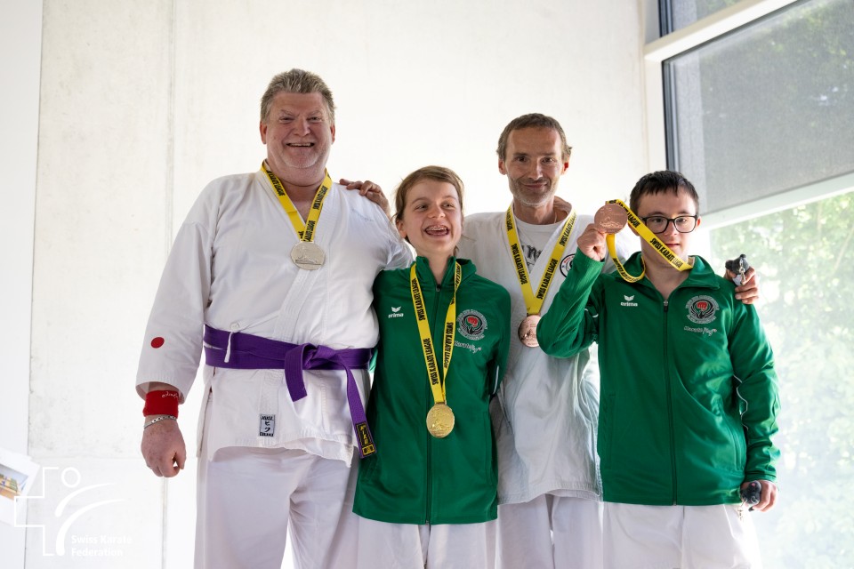 Vier lächelnde Sportler posieren gemeinsam mit Medaillen um den Hals. Einer trägt einen weißen Karateanzug mit lila Gürtel, die anderen drei grüne Sportjacken. Sie stehen vor einer weißen Wand und einem großen Fenster.