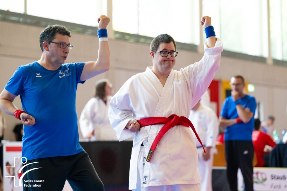 Zwei Männer, einer trägt einen weissen Karate-Gi mit rotem Gürtel, der andere ein blaues Shirt, führen in einer hellen Turnhalle nebeneinander Karateübungen aus. In der Ecke ist das Logo des Schweizerischen Karateverbands zu sehen.