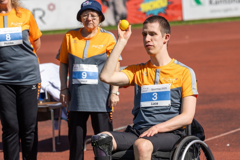 Ein junger männlicher Sportler im Rollstuhl bereitet sich darauf vor, bei einer Sportveranstaltung einen gelben Ball zu werfen. Er trägt ein orange-graues Trikot, und andere Teilnehmer in ähnlichen Uniformen stehen im Hintergrund.