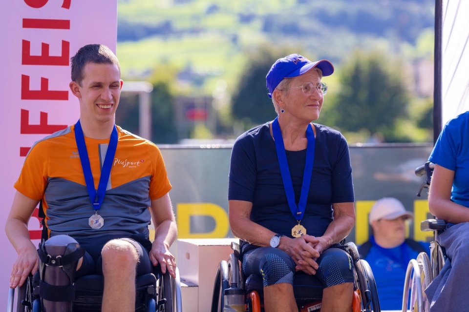 Zwei Sportler in Rollstühlen, beide mit Medaillen um den Hals, sitzen im Freien nebeneinander. Sie lächeln und scheinen bei einer Sportveranstaltung zu sein. Im Hintergrund sind Grünflächen und andere Teilnehmer zu sehen.