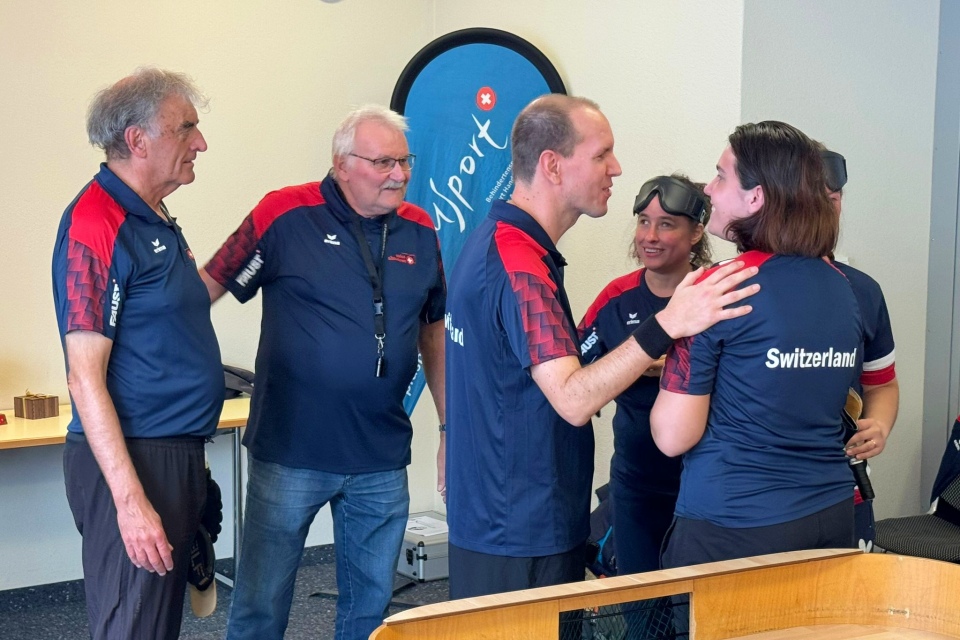 Eine Gruppe von fünf Personen in passenden marineblauen und roten Sporthemden, auf deren Rücken die Schweiz aufgedruckt ist, steht in einem Haus und unterhält sich lächelnd. Eine Person scheint eine andere zu trösten. Im Vordergrund steht ein runder Tisch.