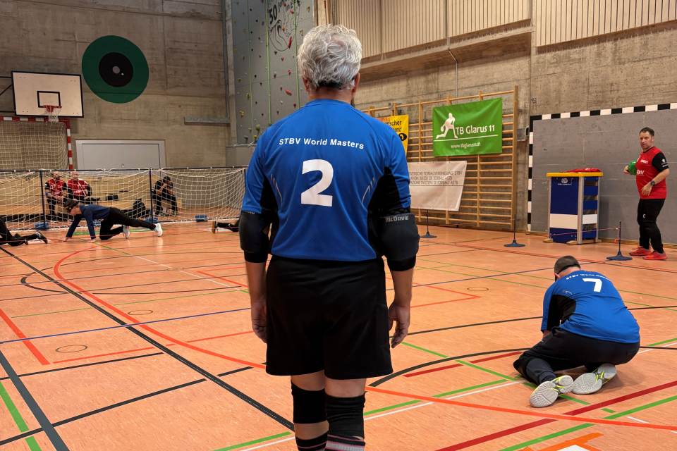 Ein Mann, der ein blaues Trikot mit der Aufschrift STBV World Masters und der Nummer 2 trägt, steht auf einem Hallenplatz und blickt auf ein Tor. Andere Spieler, ebenfalls in Blau, knien oder sitzen auf dem Platz. Es scheint ein Spiel im Gange zu sein.