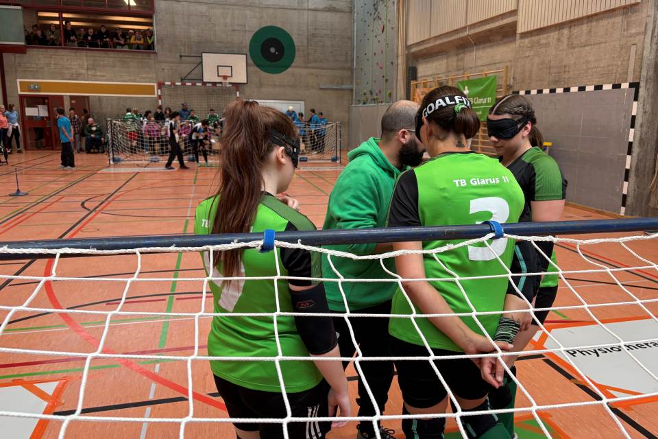 Vier Torballspieler in grünen Trikots, einige mit Augenmasken, stehen mit einem Trainer in der Nähe des Netzes in einer Sporthalle zusammen. Im Hintergrund sind andere Mannschaften und Zuschauer zu sehen.