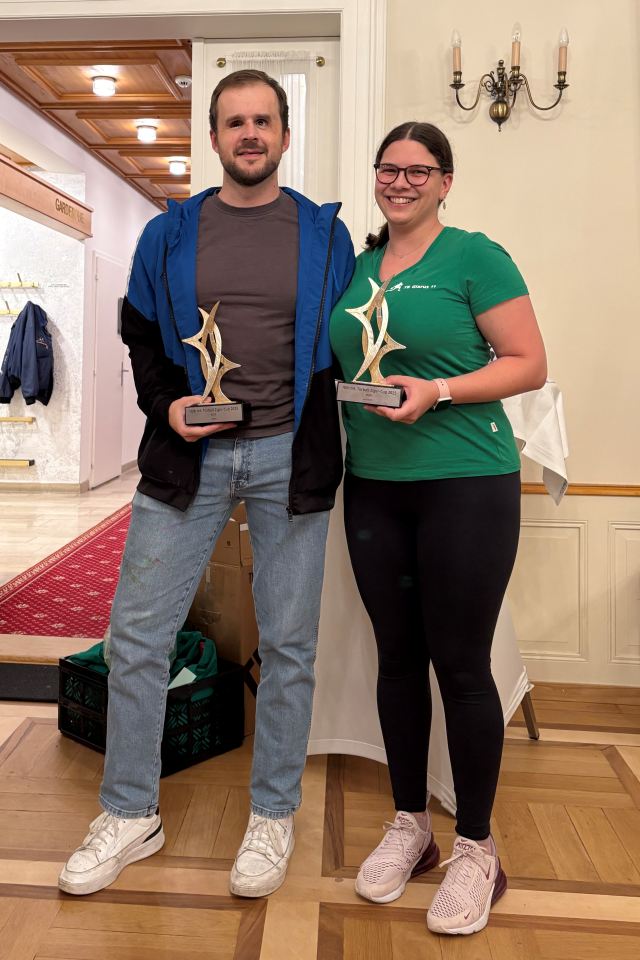 Zwei Personen stehen in einem Haus, lächeln und halten zusammengehörige Trophäen in abstrakter Form. Der Mann trägt eine blaue Jacke und Jeans, und die Frau mit Brille und grünem Hemd steht neben ihm. Sie scheinen auf einer Preisverleihung zu sein.