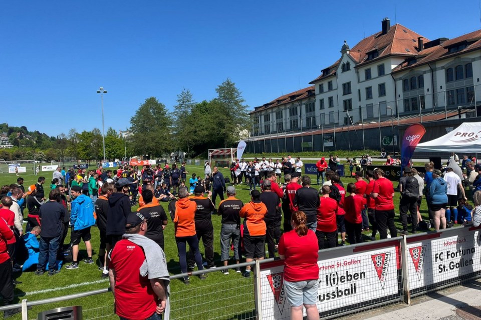 An einem sonnigen Tag versammelt sich eine große Gruppe von Menschen auf einem Fußballplatz in der Nähe eines Stadiongebäudes. Einige tragen Mannschaftsuniformen, andere schauen zu, inmitten von Bannern und Veranstaltungsfahnen.