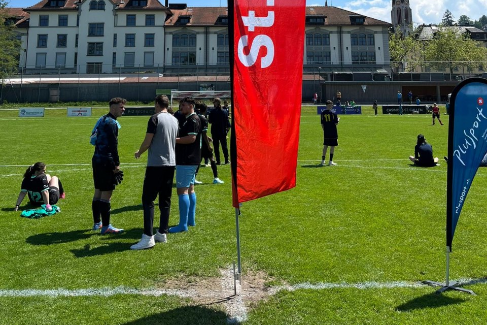 Eine Gruppe Fußballspieler steht neben einer roten Fahne auf einem sonnigen Feld, unterhält sich und ruht sich aus. Einige Spieler sitzen auf dem Rasen, andere stehen. Im Hintergrund sind Gebäude und eine Kirche zu sehen.