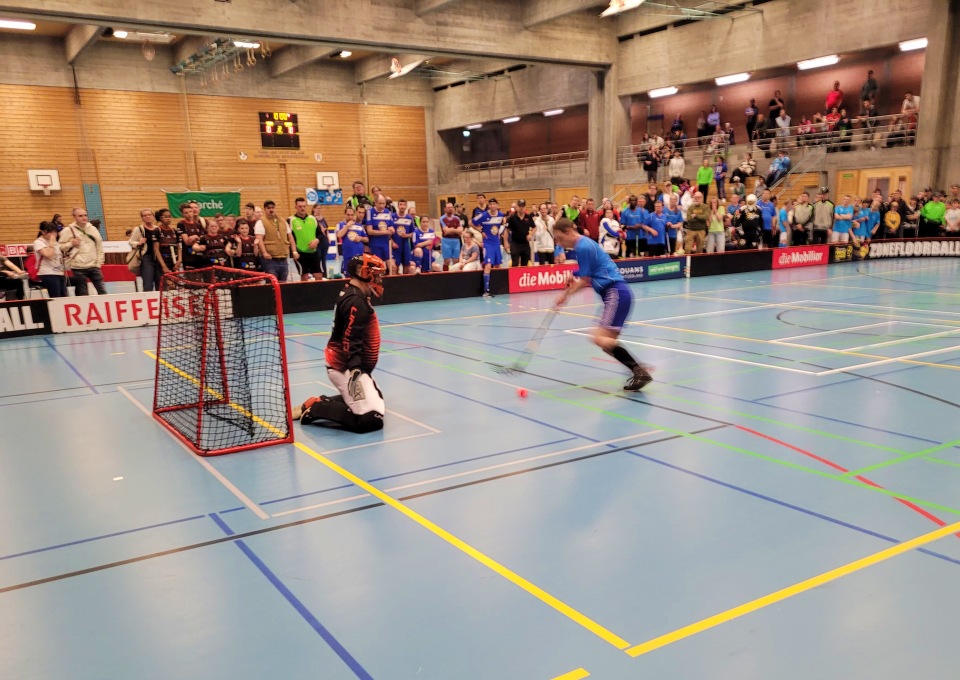 Ein Unihockeyspieler in Blau schießt aufs Tor gegen einen knienden Torwart in Schwarz und Rot. Zuschauer auf den Tribünen und entlang des Spielfelds verfolgen das Geschehen in einer hell erleuchteten Sporthalle.