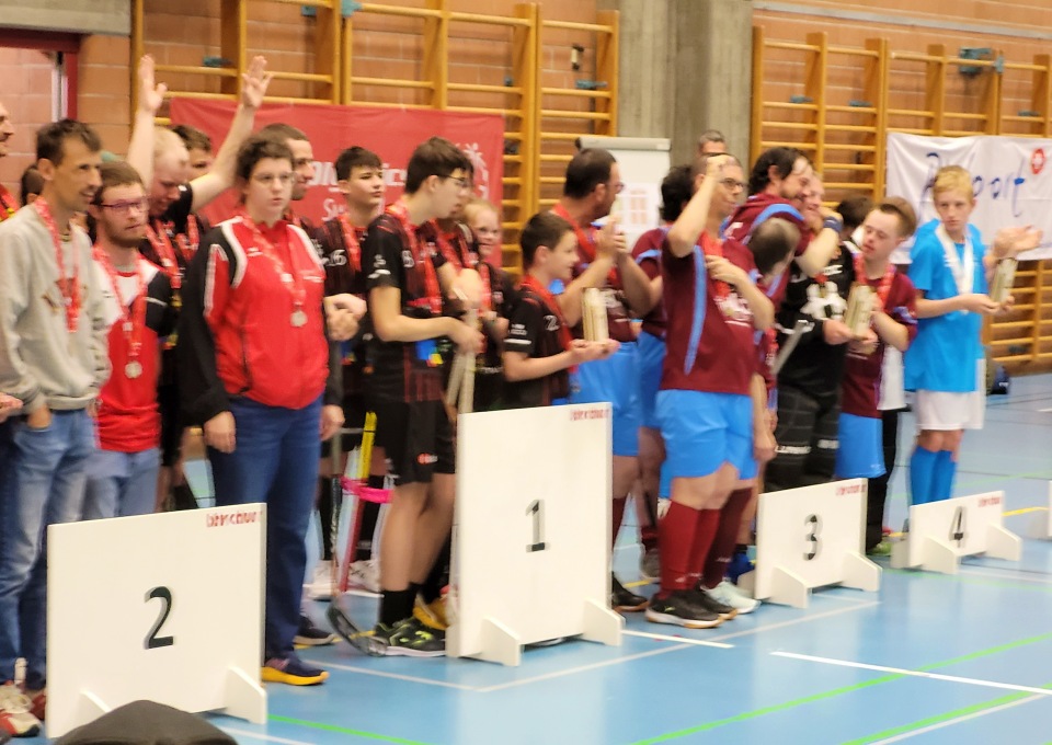 Eine Gruppe von Athleten steht mit Medaillen um den Hals auf einem Podium und feiert bei einer Hallensportveranstaltung. Das Podium ist mit den Nummern 1, 2 und 3 für verschiedene Positionen gekennzeichnet.