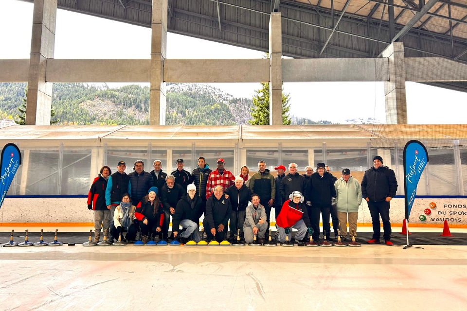 Eine Gruppe von etwa 25 Personen posiert gemeinsam in einer Eishalle, einige tragen Helme. Draußen hinter ihnen sind Berge und Bäume zu sehen. Zwei blaue Banner und ein Schild sind auf beiden Seiten der Gruppe angebracht.