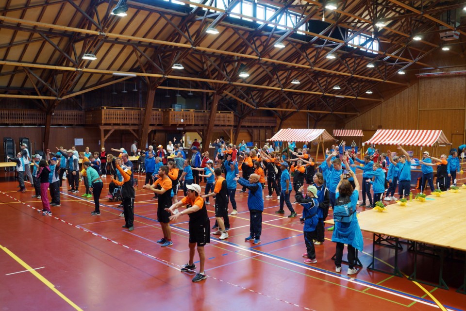 Eine große Gruppe von Menschen in farbenfroher Sportkleidung nimmt an einer Gruppenübungssitzung in einer geräumigen Holzturnhalle mit hoher Decke und Marktständen im Hintergrund teil.