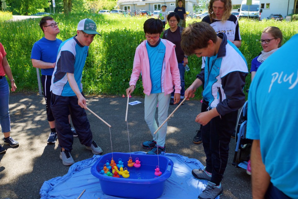 Eine Gruppe von Menschen steht um eine blaue Plastikwanne mit Wasser und Spielzeugenten herum und fängt die Enten mit Angelruten. Sie scheinen an einem sonnigen Tag draußen zu sein und einer lustigen Aktivität nachzugehen.