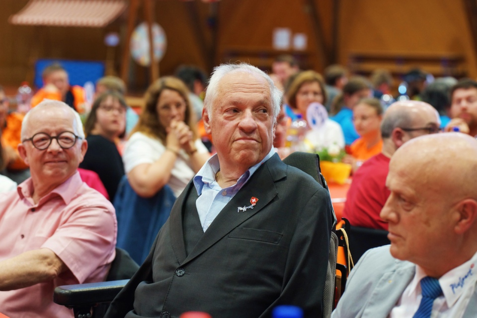 Ein älterer Mann im Anzug sitzt während einer Veranstaltung an einem Tisch, umgeben von anderen Teilnehmern. Im Hintergrund unterhalten sich die Menschen, und man sieht farbenfrohe Dekorationen.