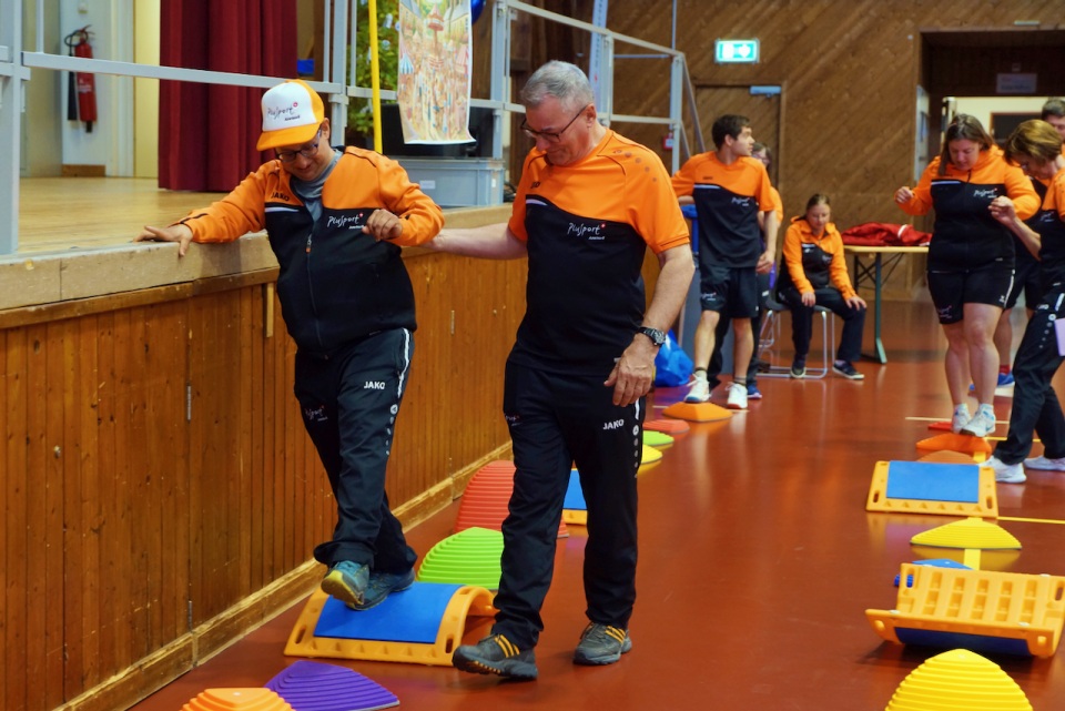 Zwei Erwachsene in passender orange-schwarzer Sportbekleidung balancieren in einer Turnhalle auf bunten Trittsteinen aus Plastik, wobei einer dem anderen hilft. Im Hintergrund sind mehrere Personen in ähnlicher Kleidung zu sehen, die dieselbe Aktivität ausüben.